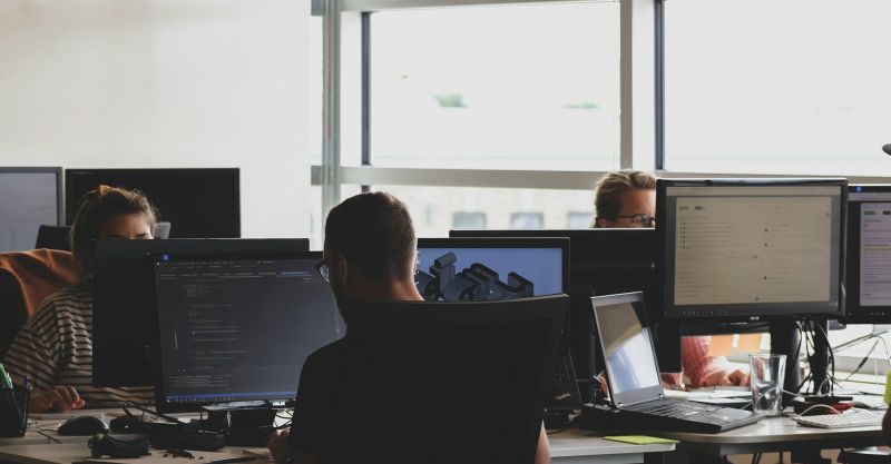 Employees in an open office space sitting on chairs in front of computer monitors.